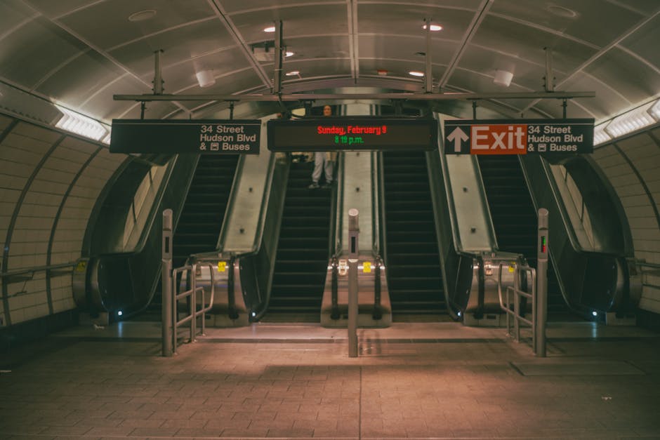 A person dressed in an orange coat and carrying a red bag is walking up a metal staircase inside a modern building, with the staircase featuring black treads and metal railings on both sides. Overhead