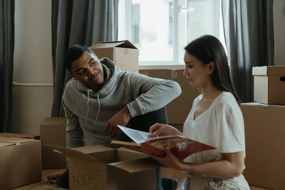 A young man and woman inside a room during a home relocation process, surrounded by cardboard boxes of various sizes, some labeled with contents such as 'BOOKS', indicating packing and moving preparations. The man, wearing a grey hoodie, is seated on the floor beside a stack of boxes, looking attentively at the woman with a slight smile. The woman, dressed in a white top, is holding an open book or document, focusing on it as part of the packing or organizing activity. The room features a large window with natural light coming through, partially covered by dark curtains. Cardboard boxes are placed on the floor and on a windowsill, with some packed and others open for packing or unpacking. There is no visible furniture or appliances; the environment appears to be an active packing area during a furniture transport or home move. The scene underscores aspects of packing, organizing, and preparing for a residential move, supported by the presence of packing materials and the careful handling of items during the process, which may involve moving services from Man with Van New Southgate.
