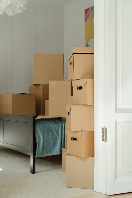 A room filled with stacked cardboard boxes of varying sizes, some with handles, placed on a bed with a blue bedspread and partially extending into a doorway. The boxes appear to be prepared for a house removal or furniture transport process, with additional packing materials possibly inside. The area is well-lit, with a white wall featuring a colorful abstract painting in the background. Inside the room, furniture such as a grey painted bed frame is visible, indicating a residential setting. The boxes are positioned both on the bed and directly on the floor, with some partially blocking the doorway. This scene illustrates the packing and loading process typical of home relocation services by Man with Van New Southgate, demonstrating organized preparation for furniture transport and moving logistics.