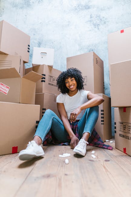 A young man and woman inside a room during a home relocation process, surrounded by cardboard boxes of various sizes, some labeled with contents such as 'BOOKS', indicating packing and moving preparations. The man, wearing a grey hoodie, is seated on the floor beside a stack of boxes, looking attentively at the woman with a slight smile. The woman, dressed in a white top, is holding an open book or document, focusing on it as part of the packing or organizing activity. The room features a large window with natural light coming through, partially covered by dark curtains. Cardboard boxes are placed on the floor and on a windowsill, with some packed and others open for packing or unpacking. There is no visible furniture or appliances; the environment appears to be an active packing area during a furniture transport or home move. The scene underscores aspects of packing, organizing, and preparing for a residential move, supported by the presence of packing materials and the careful handling of items during the process, which may involve moving services from Man with Van New Southgate.