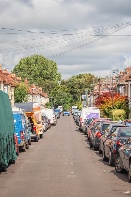 A street scene featuring a narrow residential road leading to a property with a brick and white facade. The pavement on both sides has trees and pedestrians walking along the walkway. Two buses are pa